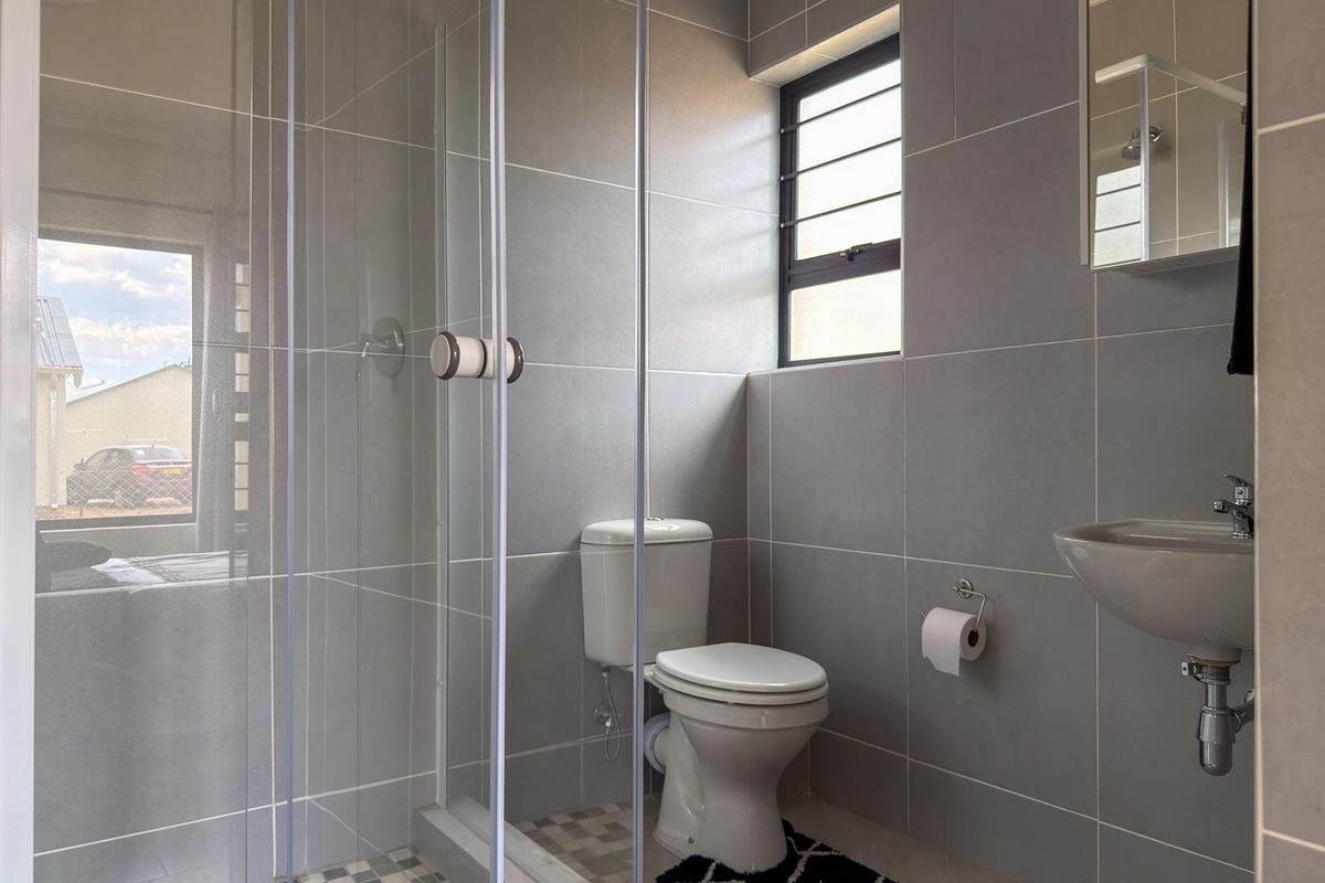 Modern bathroom featuring a glass-enclosed shower, wall-mounted basin, and toilet, finished with neutral grey wall tiles and a window for natural light.