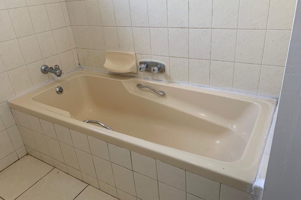 A dated bathroom featuring a built-in bathtub with chrome fixtures and ceramic wall tiling, requiring cosmetic updates to modernize the space.