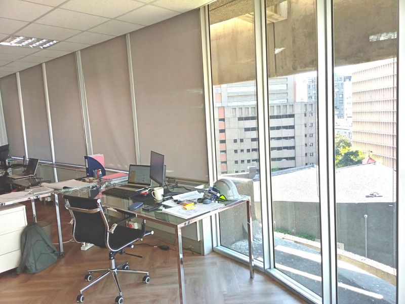 A bright office space featuring parquet flooring, suspended ceilings, and expansive floor-to-ceiling windows offering an urban city view.