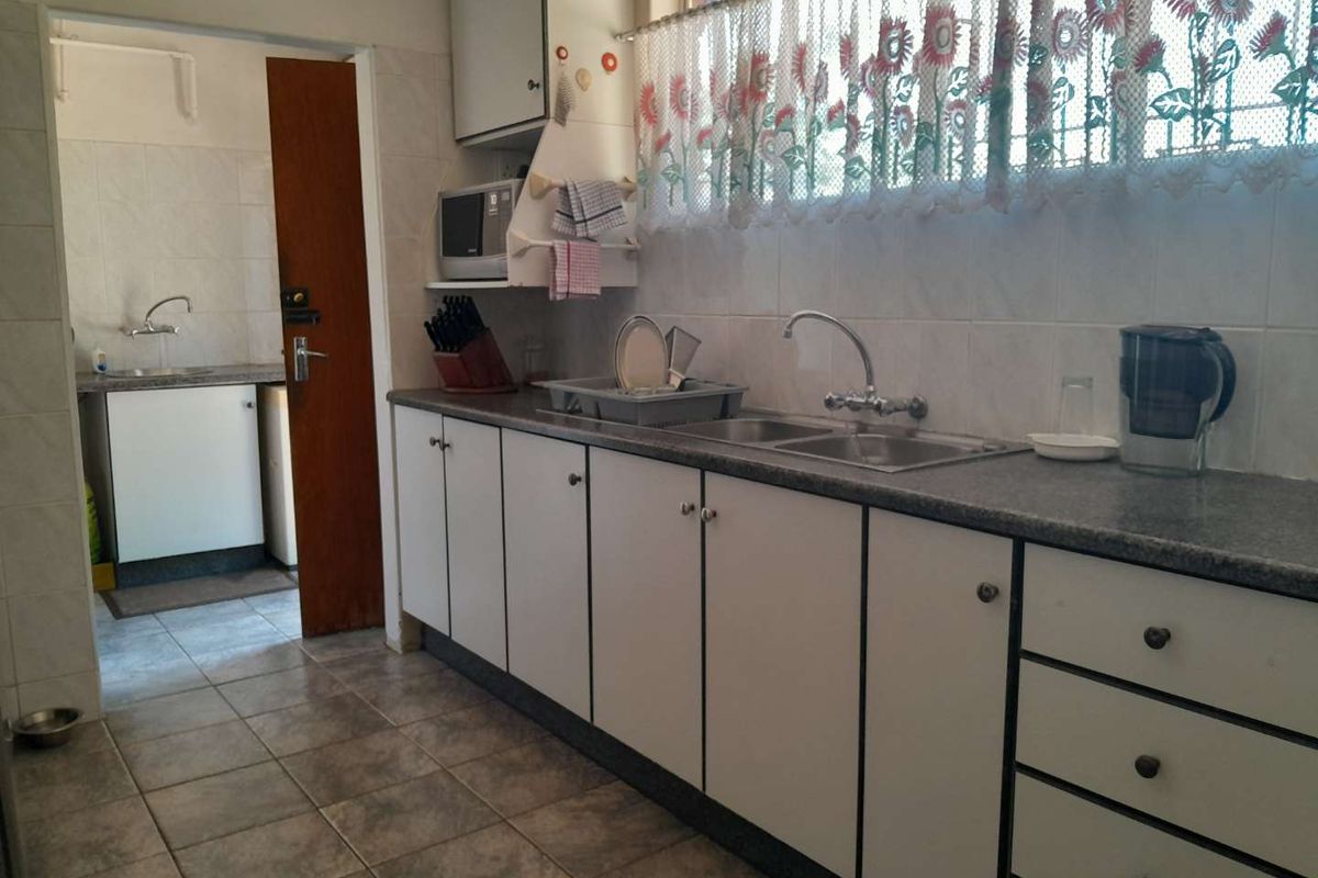 A functional kitchen featuring white cabinetry, a double stainless steel sink, and tiled flooring, with an adjacent scullery area visible through the doorway.