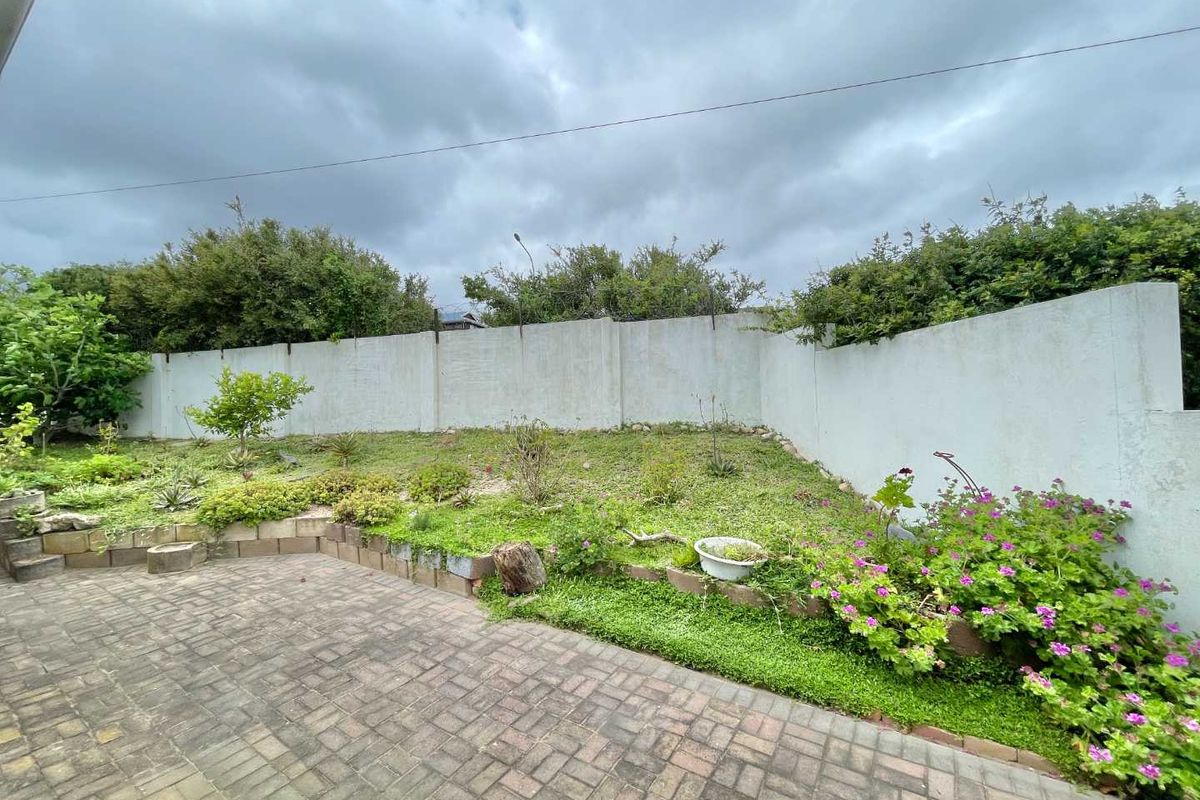 A rear garden area featuring a paved patio, tiered landscaping with a retaining wall, and a high boundary wall.