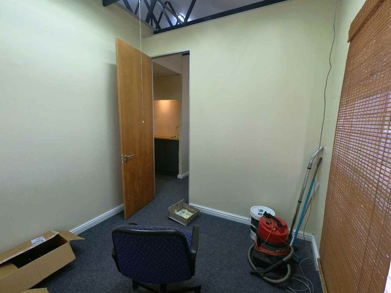 A vacant office space featuring exposed roof trusses, carpeted flooring, and a timber door leading to an adjacent utility area.