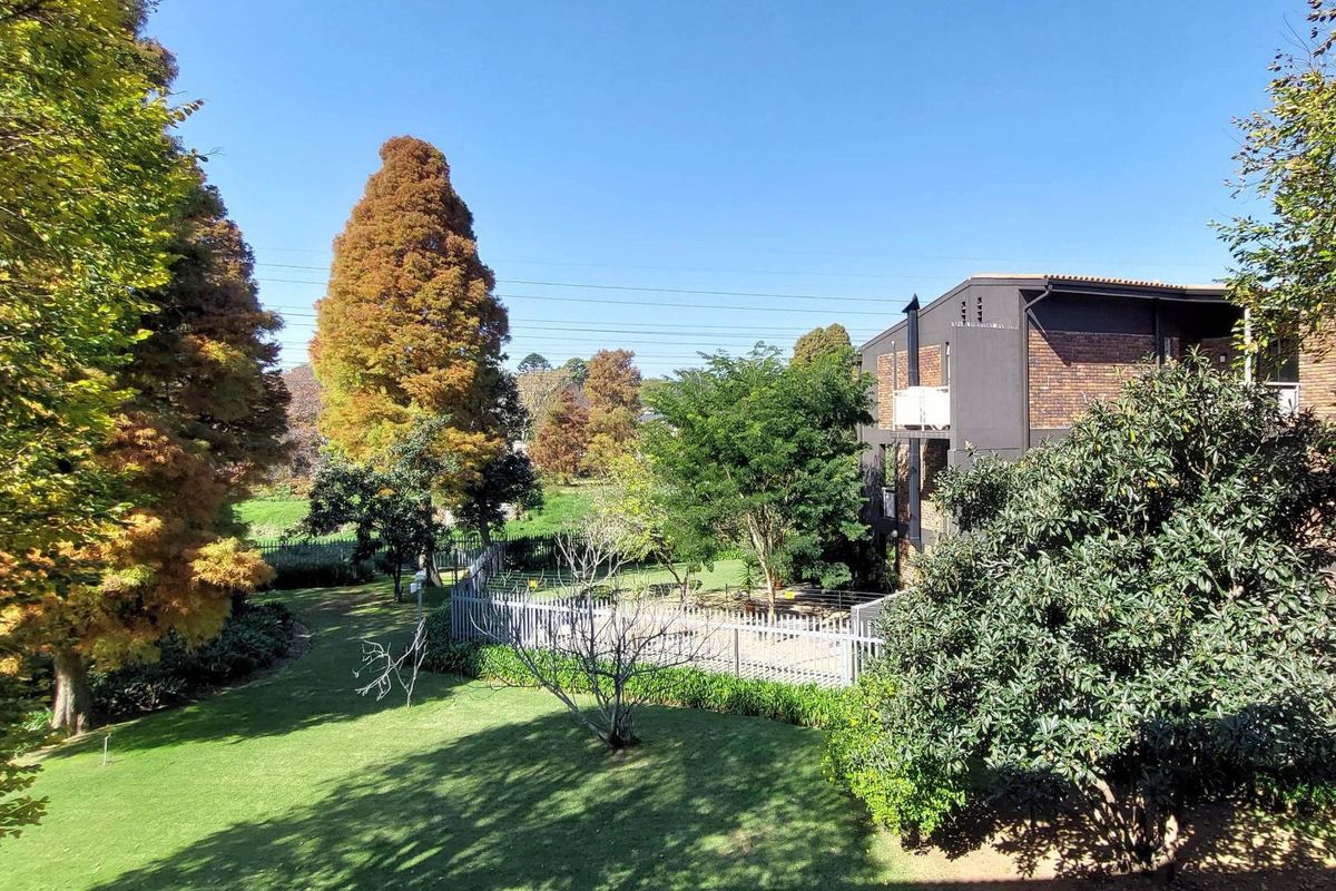 View of the apartment building exterior featuring a brick facade, private balcony, and well-maintained communal garden area with mature trees.
