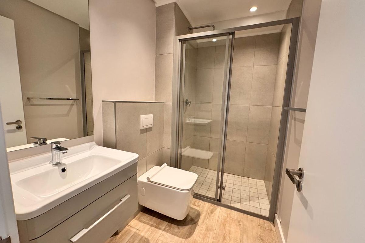 Modern bathroom featuring a wall-mounted toilet, vanity basin, and a glass-enclosed shower with neutral tiling and wood-look flooring.