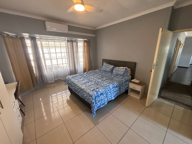 Spacious bedroom featuring tiled flooring, a ceiling fan, and a wall-mounted air conditioning unit, with large windows providing natural light.