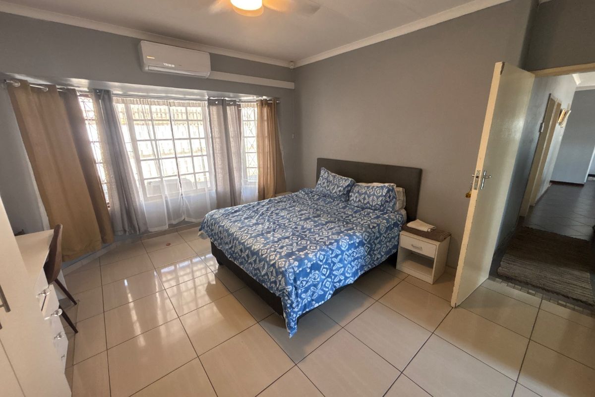 Spacious bedroom featuring tiled flooring, a ceiling fan, and a wall-mounted air conditioning unit, with large windows providing natural light.