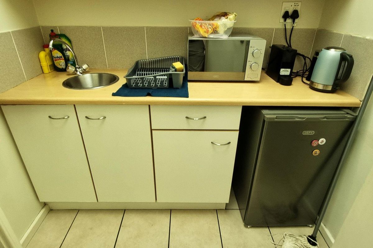 A compact office kitchenette featuring built-in cabinetry, a stainless steel sink, tiled backsplash, and space for a small under-counter refrigerator.