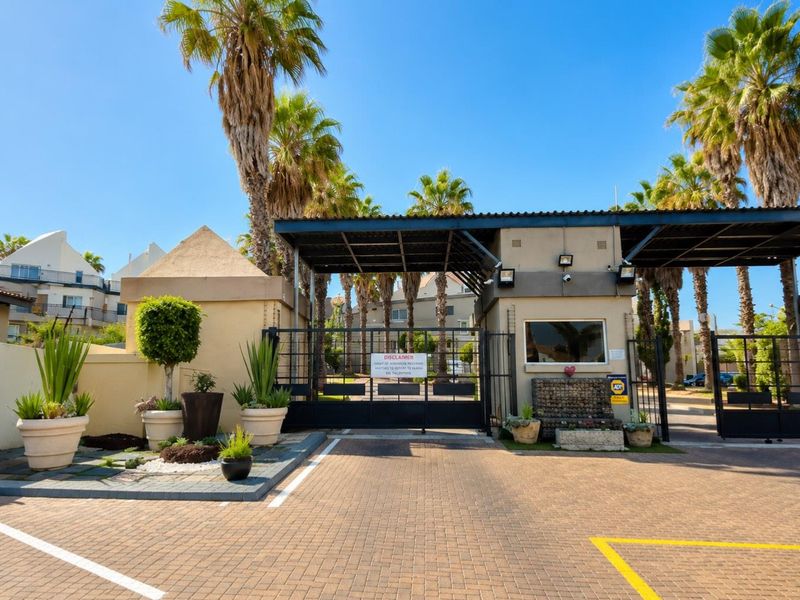 A secure gated entrance to the residential complex featuring a guard house, paved driveway, and established palm trees.