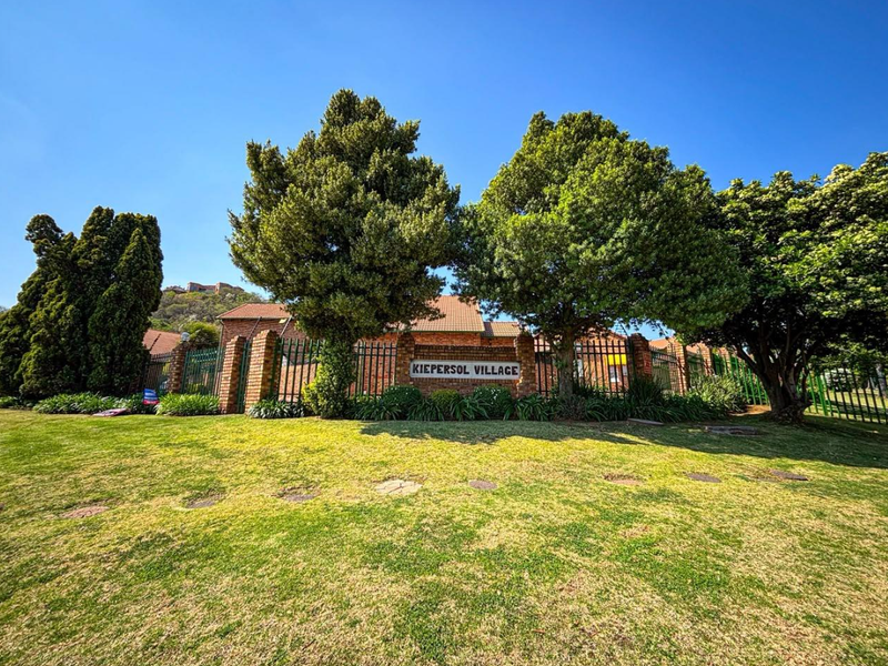 Front view of Kiepersol Village townhouse complex featuring a brick boundary wall, security fencing, and well-maintained garden area.