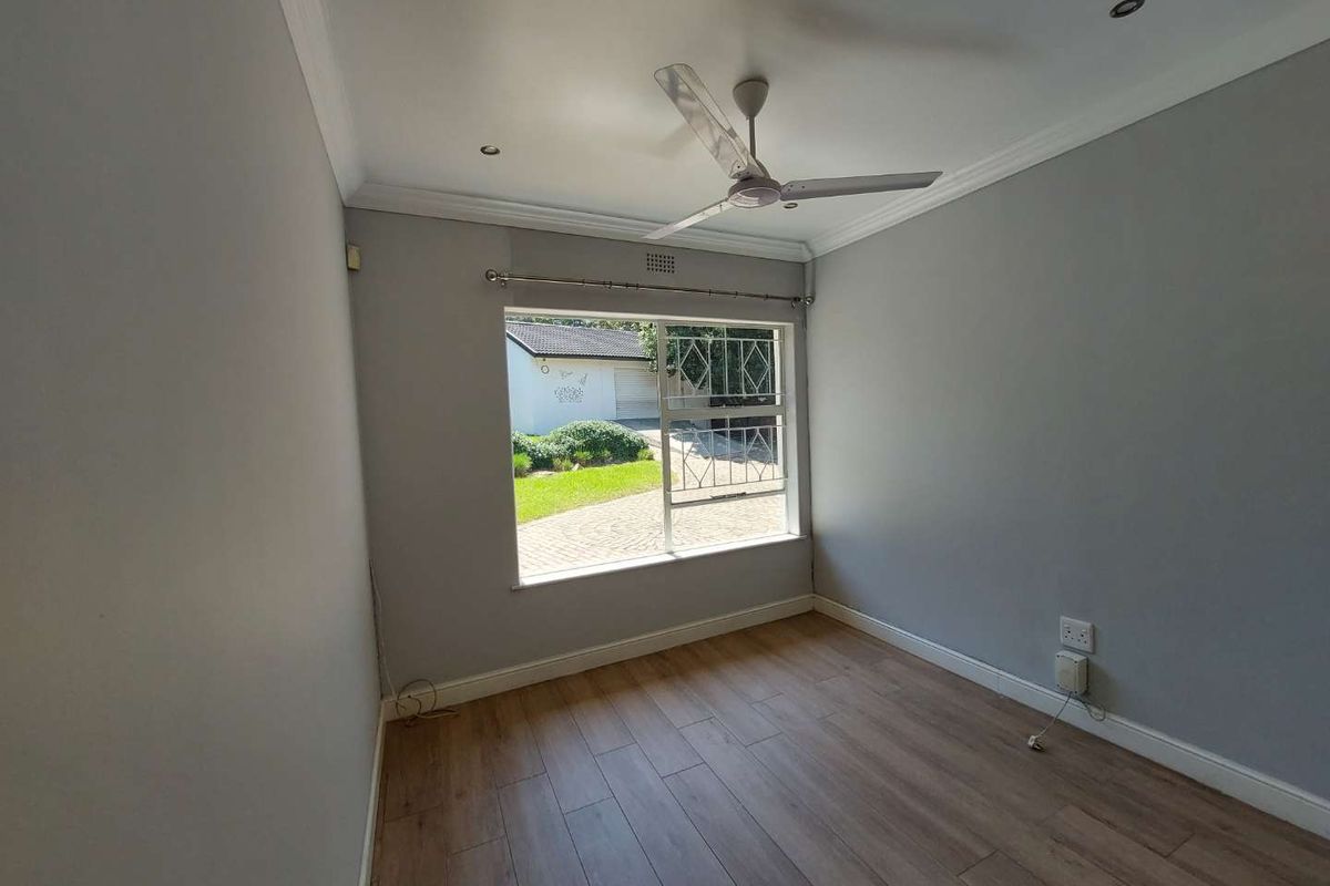 A bright, empty bedroom featuring modern laminate flooring, a ceiling fan, and a window with security bars overlooking the front garden.