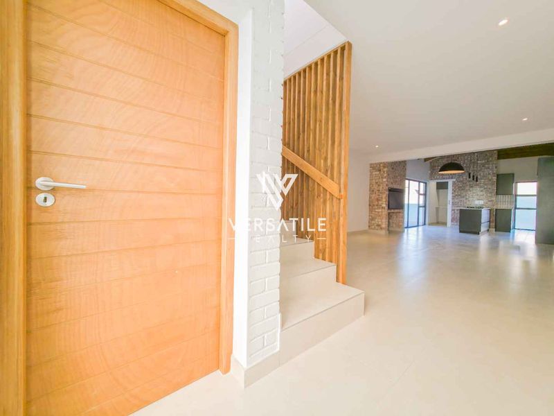 Spacious open-plan living area featuring a modern timber staircase with slatted balustrade, exposed brick accents, and neutral tiled flooring throughout.