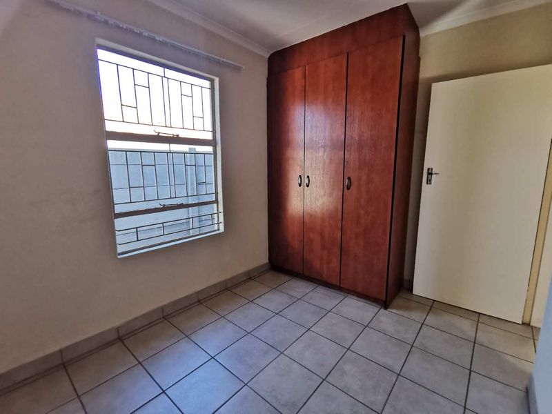 A clean, empty bedroom featuring tiled flooring, a built-in wooden wardrobe, and a window fitted with security bars.