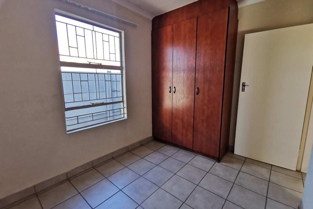 A clean, empty bedroom featuring tiled flooring, a built-in wooden wardrobe, and a window fitted with security bars.