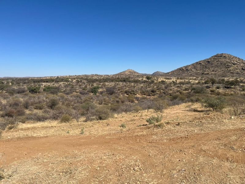 A wide-angle view of the natural landscape featuring arid terrain, native shrubs, and distant mountain ranges under a clear blue sky.
