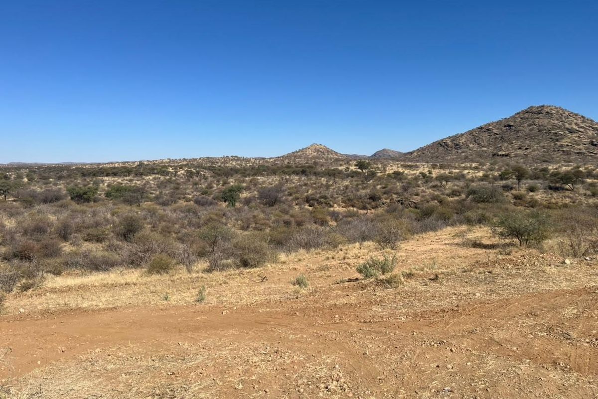 A wide-angle view of the natural landscape featuring arid terrain, native shrubs, and distant mountain ranges under a clear blue sky.