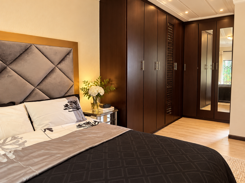 A well-appointed bedroom featuring extensive dark wood built-in cupboards with integrated mirrors, recessed ceiling lighting, and light-toned laminate flooring.