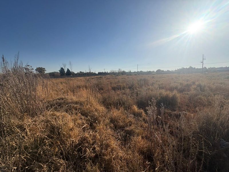 A wide-angle view of a vacant residential plot featuring dry grass and perimeter fencing under a clear blue sky.
