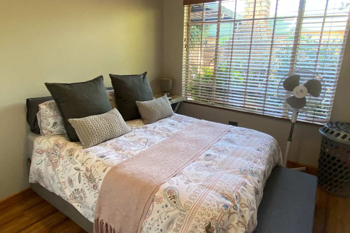 A cozy bedroom featuring warm-toned wooden flooring and large windows with fitted blinds, providing natural light and a view of the exterior.