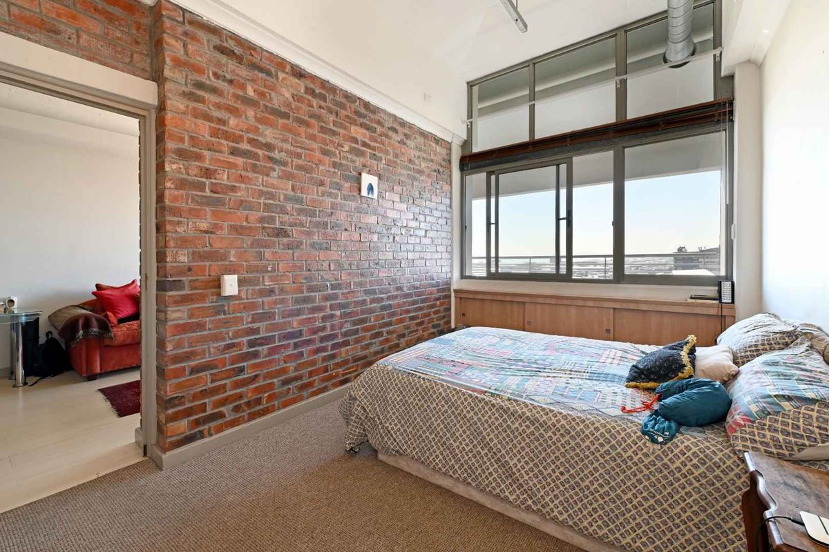 A bedroom featuring an exposed brick feature wall, large windows with natural light, and carpeted flooring, with an adjacent room visible through the doorway.