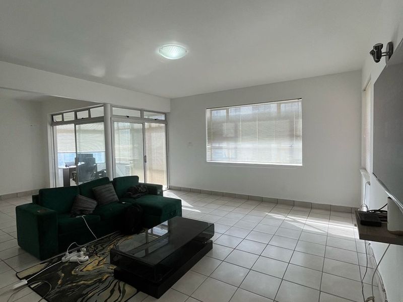A spacious living room featuring neutral tiled flooring, large windows with venetian blinds, and sliding glass doors leading to an adjacent enclosed balcony area.