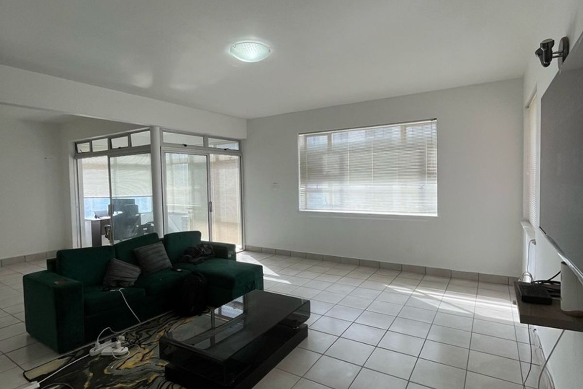 A spacious living room featuring neutral tiled flooring, large windows with venetian blinds, and sliding glass doors leading to an adjacent enclosed balcony area.