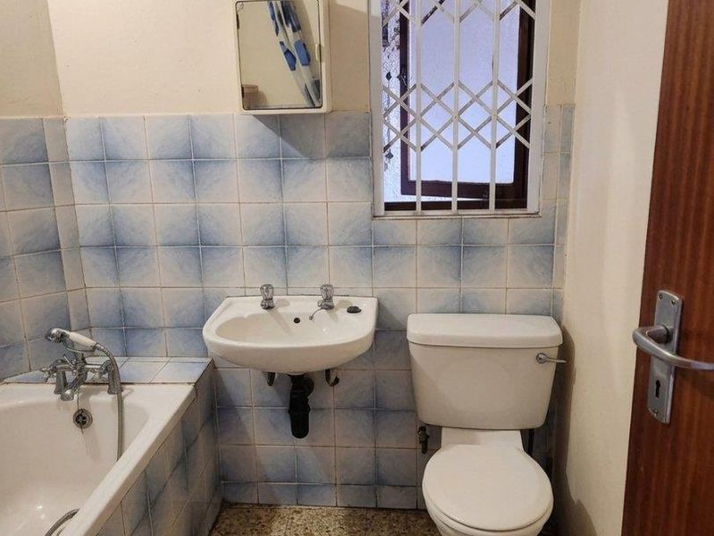 A compact bathroom featuring a bathtub, wall-mounted basin, and toilet, with dated tiling and a security-gated window.
