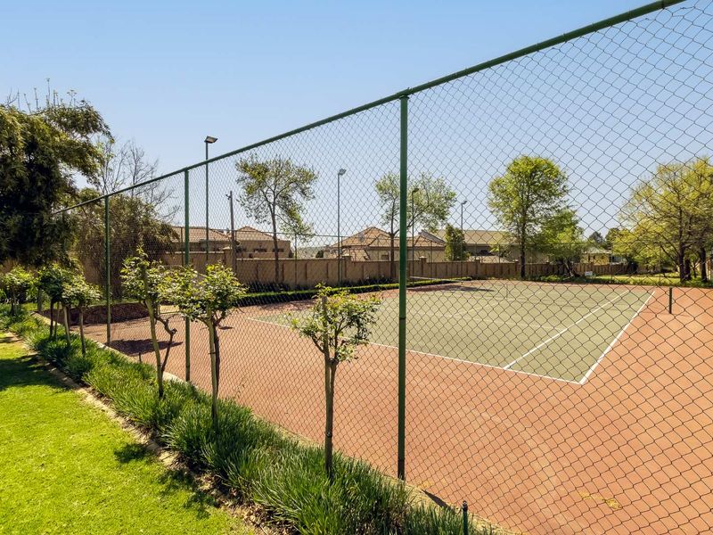 A well-maintained outdoor tennis court featuring a durable surface, perimeter chain link fencing, and integrated floodlighting within a landscaped residential complex.