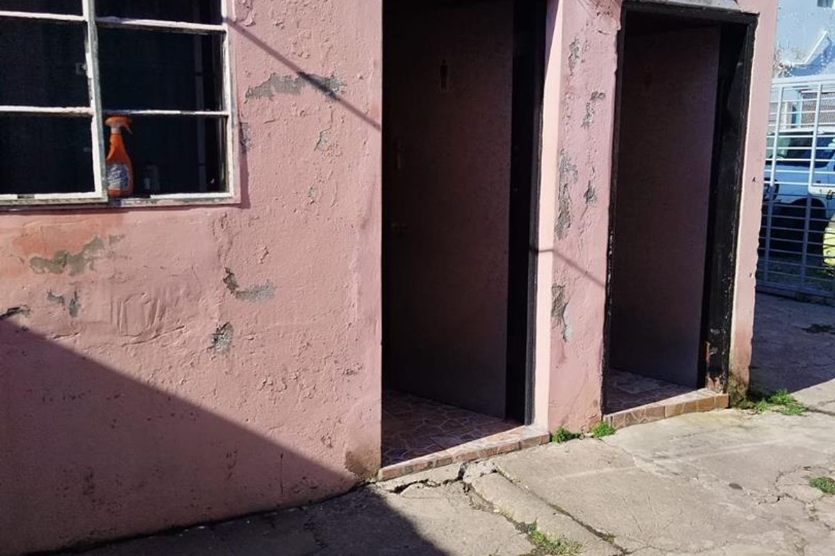Exterior view of a utility building featuring two doorways and a metal-framed window, showing significant signs of wear and structural weathering.