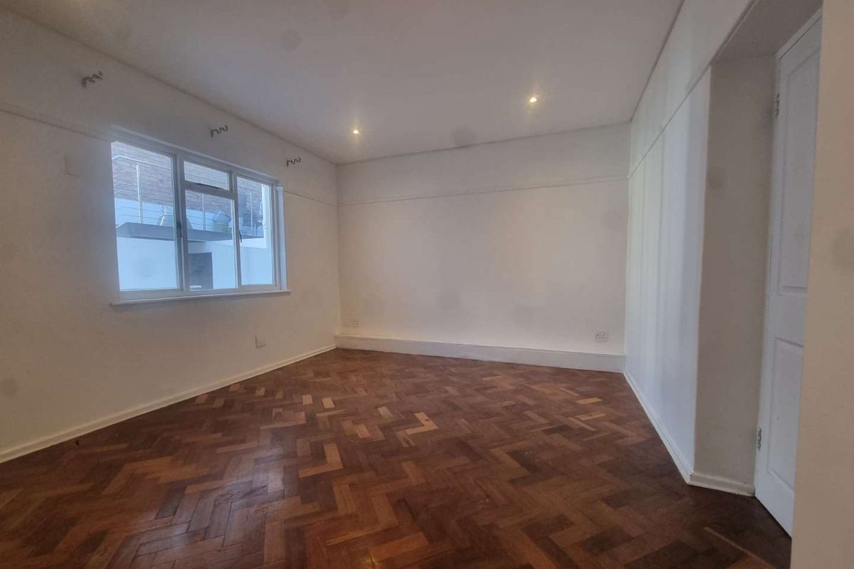 A bright, empty living room featuring classic herringbone parquet flooring, recessed ceiling lighting, and a large window providing natural light.