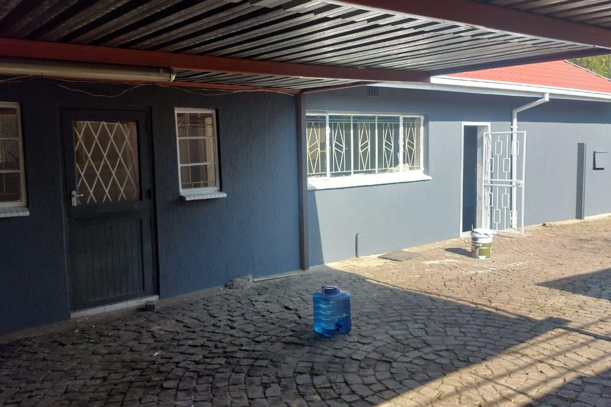 Rear exterior view featuring a covered carport area, cobblestone paving, and secure access points with metal security gates and window bars.