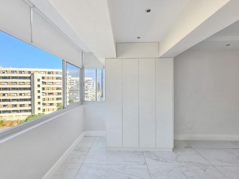 Bright and modern bedroom featuring sleek built-in cupboards, marble-look floor tiles, recessed lighting, and large windows offering an urban view.