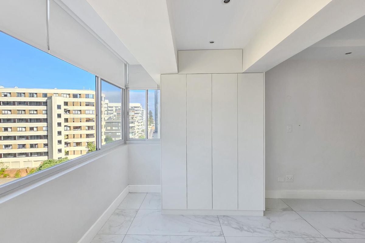 Bright and modern bedroom featuring sleek built-in cupboards, marble-look floor tiles, recessed lighting, and large windows offering an urban view.