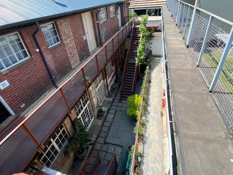 An elevated view of an industrial-style apartment complex courtyard featuring exposed brickwork, a metal staircase, and a concrete walkway with mesh railings.