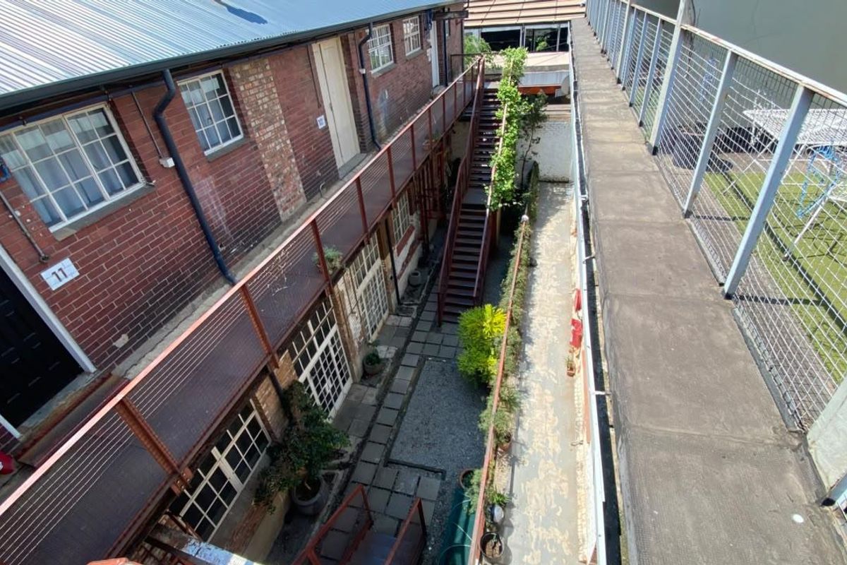An elevated view of an industrial-style apartment complex courtyard featuring exposed brickwork, a metal staircase, and a concrete walkway with mesh railings.