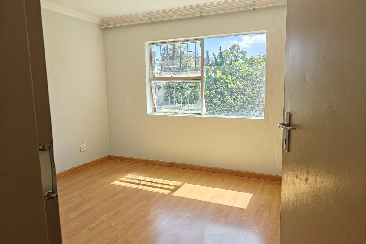 A bright, empty bedroom featuring light-toned laminate flooring, a large window with security bars, and neutral wall finishes.