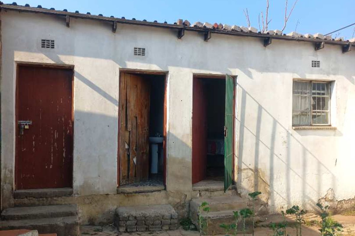 Rear exterior view of a single-story dwelling featuring multiple entry doors, concrete steps, and a paved area requiring maintenance and exterior refurbishment.