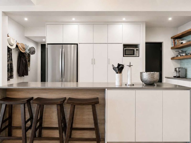 Modern kitchen featuring a central island with stone countertops, sleek white cabinetry, floating timber shelving, and a stylish subway tile backsplash.