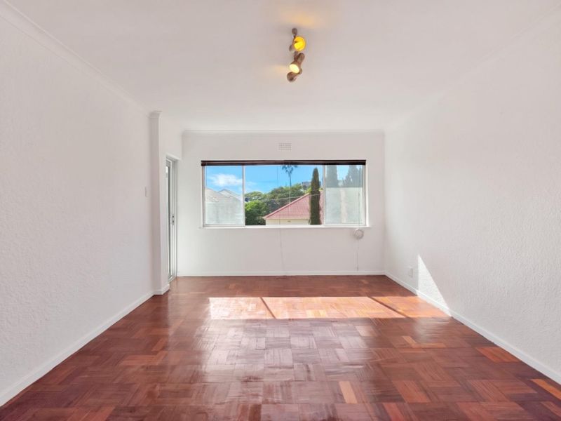 A bright, empty living room featuring polished parquet flooring, a large window with natural light, and a modern ceiling light fixture.