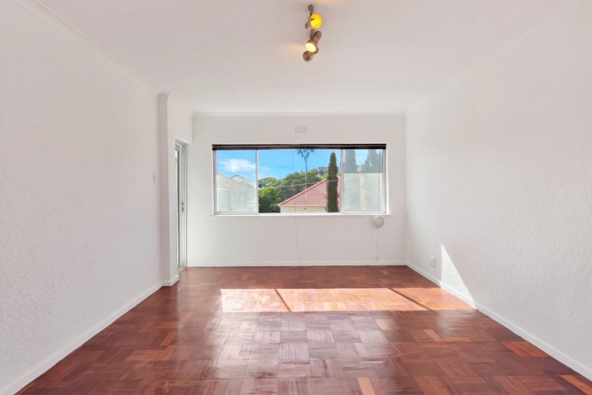 A bright, empty living room featuring polished parquet flooring, a large window with natural light, and a modern ceiling light fixture.