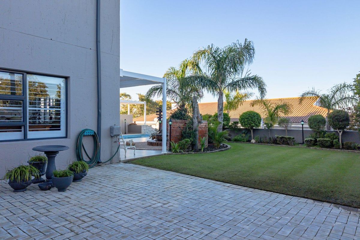 A well-maintained rear garden featuring a paved patio area, manicured lawn, established landscaping, and a view towards the swimming pool and covered patio.