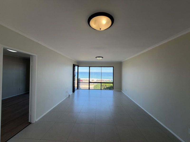 Spacious, empty living area featuring neutral tiled flooring, ceiling lighting, and a large window providing expansive ocean views.
