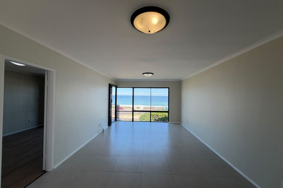 Spacious, empty living area featuring neutral tiled flooring, ceiling lighting, and a large window providing expansive ocean views.