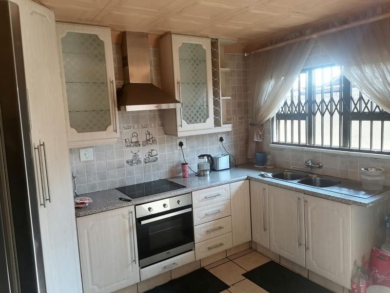 A functional kitchen featuring built-in cabinetry, a stainless steel sink, electric hob, oven, and extractor fan, with a window fitted with a security gate.