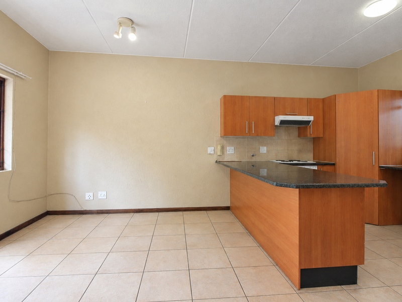 An open-plan kitchen area featuring wooden cabinetry, granite countertops, an extractor fan, and neutral tiled flooring in a clean, vacant space.