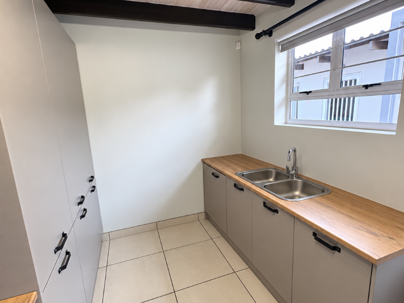 A modern, clean scullery featuring neutral cabinetry, wood-look countertops, a double sink, and exposed timber ceiling beams.