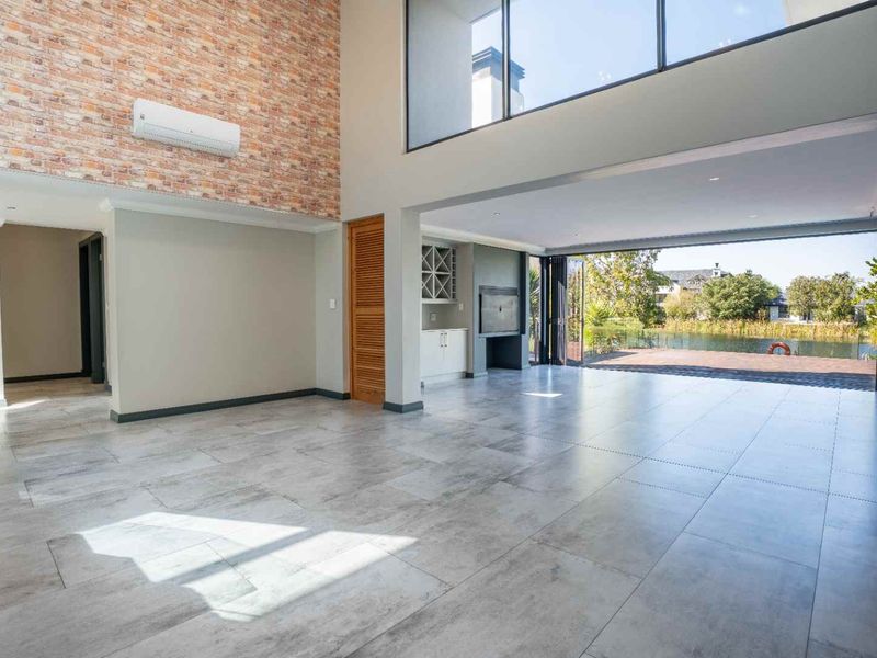 Spacious open-plan living area featuring double volume ceilings, exposed brickwork, built-in bar, fireplace, and seamless flow to the outdoor patio through stacking glass doors.