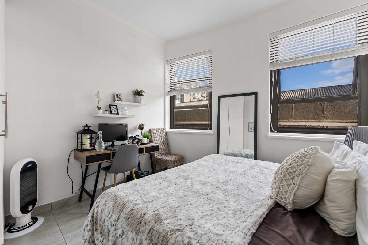 A bright bedroom featuring tiled flooring, built-in shelving, and large windows with venetian blinds, offering a clean and functional living space.