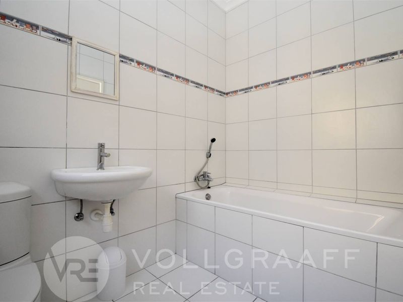 A functional bathroom featuring a built-in bathtub with handheld shower mixer, wall-mounted basin, and neutral ceramic tiling throughout.