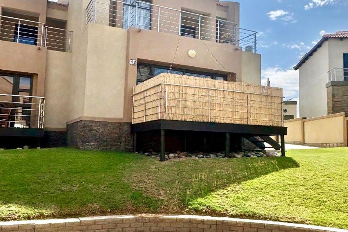 Rear view of a townhouse featuring a raised timber deck with privacy screening, stone base detailing, and a paved patio area overlooking the lawn.