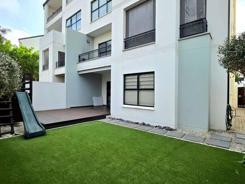 Rear view of the apartment building featuring a private garden with artificial lawn, timber decking, and a paved pathway leading to the building entrance.